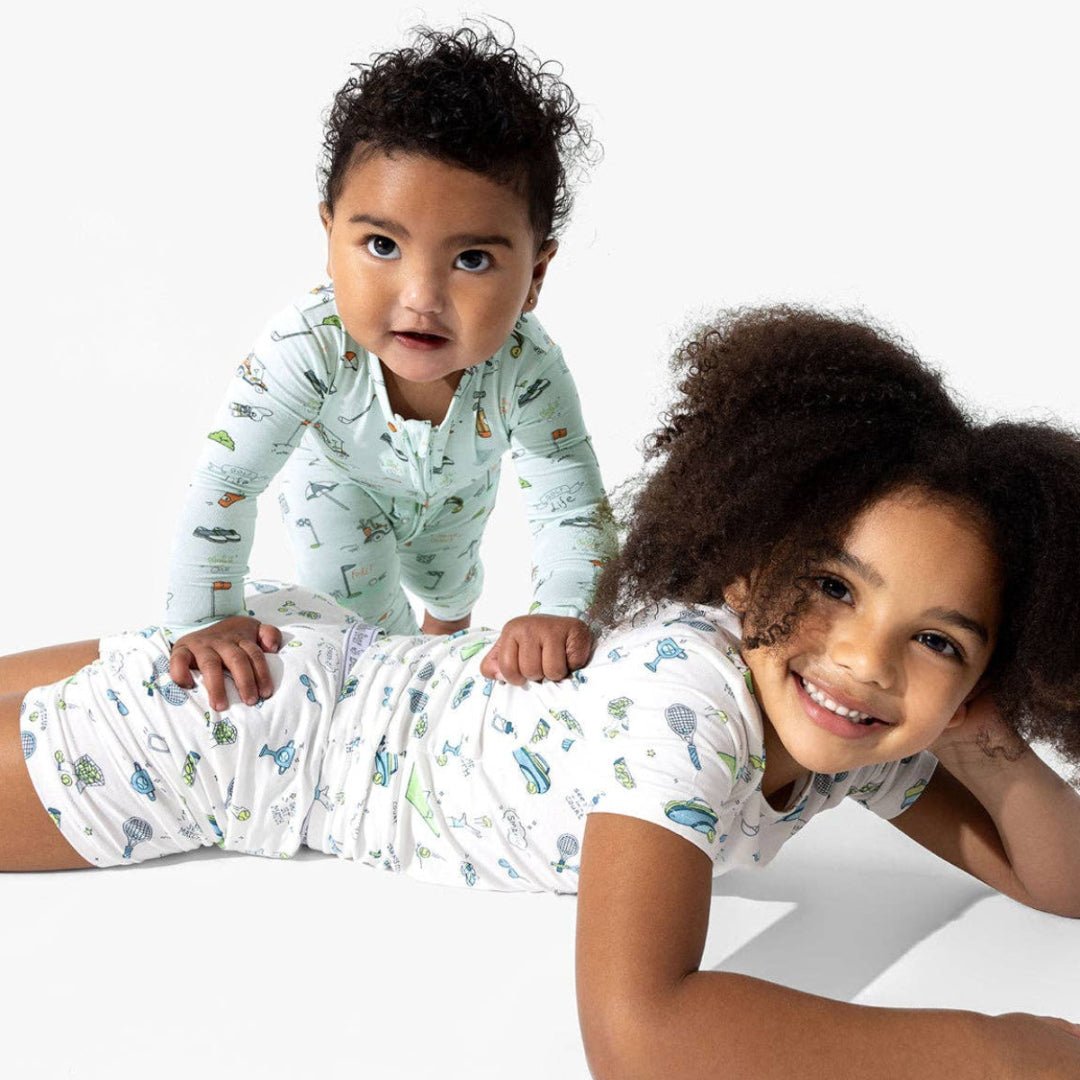 Two children in pajamas lying on a white surface. Young baby has light blue golf patterned zip up jammies and older sister has white tennis themed jammies. 
