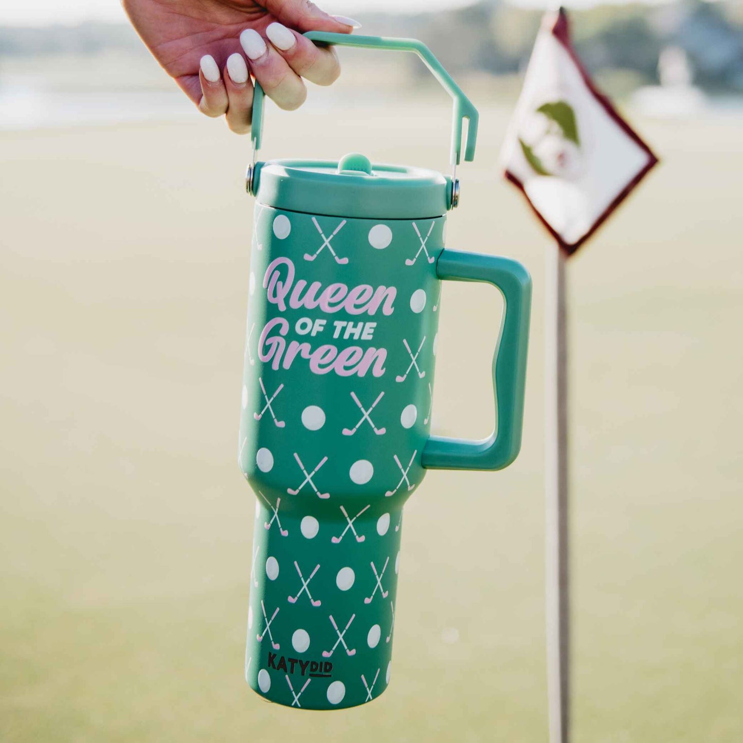 Hand holding a green “Queen of the Green” golf tumbler with handle on a golf course, with a golf flag visible in the background.