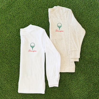 Front of two long sleeve kids’ golf t-shirts, one white and one heather beige, each featuring a small green golf ball on a tee graphic with the word “Champion” in orange script, displayed on grass background.