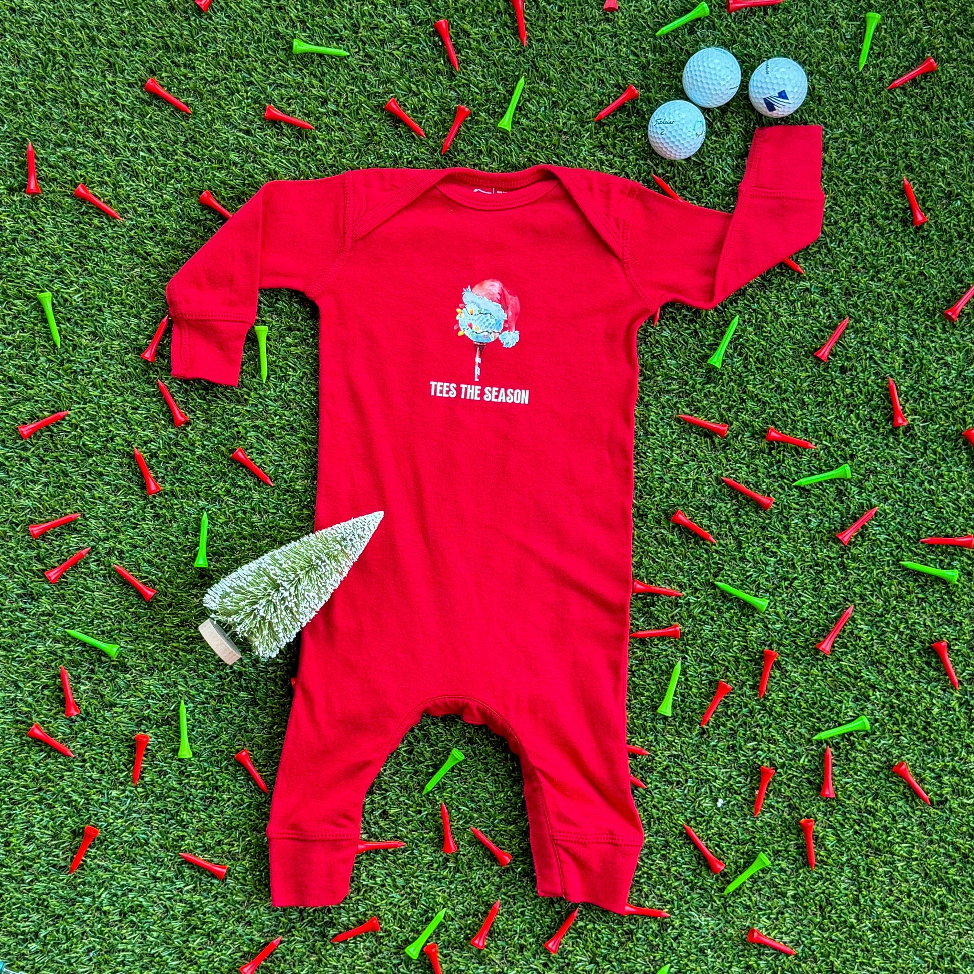 Red baby coverall with a “Tees the Season” Christmas golf graphic displayed on turf, surrounded by red and green golf tees, holiday lights, and a small bottlebrush tree.