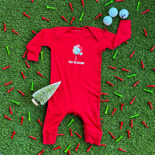 Red baby coverall with a “Tees the Season” Christmas golf graphic displayed on turf, surrounded by red and green golf tees, holiday lights, and a small bottlebrush tree.