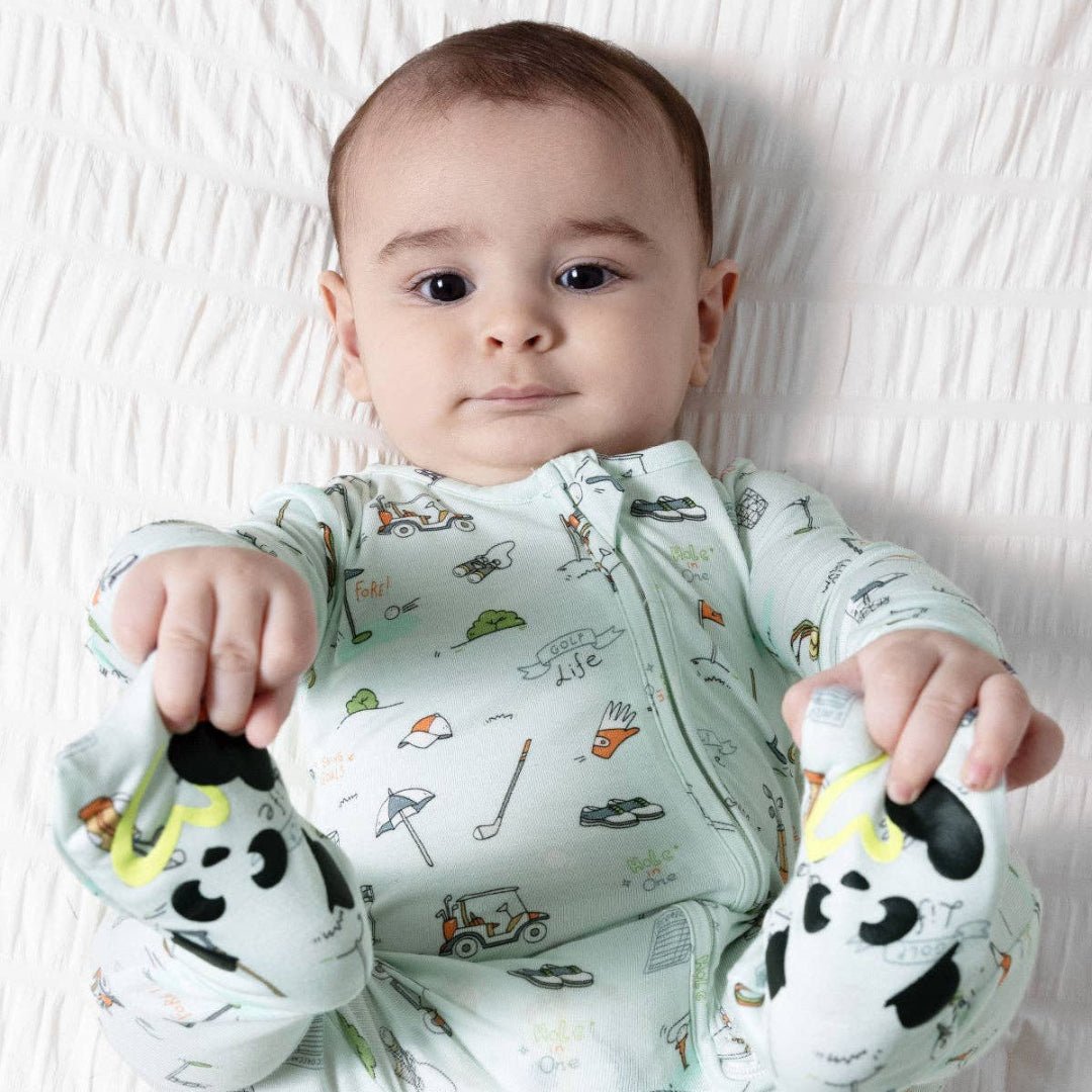 A baby lying on a white textured blanket, wearing a pale mint green long-sleeve zip-up sleeper with a colorful cartoon golf print.