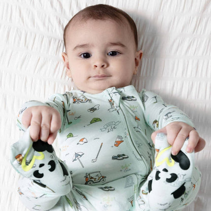 A baby lying on a white textured blanket, wearing a pale mint green long-sleeve zip-up sleeper with a colorful cartoon golf print.