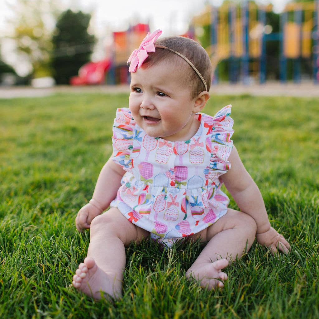Baby sitting on grass wearing a onesie with ruffle sleeves and a colorful pickleball paddle and bow pattern. 
