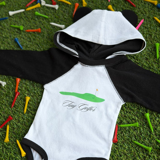 Unisex baby hooded bodysuit with black sleeves and bear ears, featuring a green golf course graphic and the words “Tiny Golfer” on the front, displayed on grass with colorful golf tees.