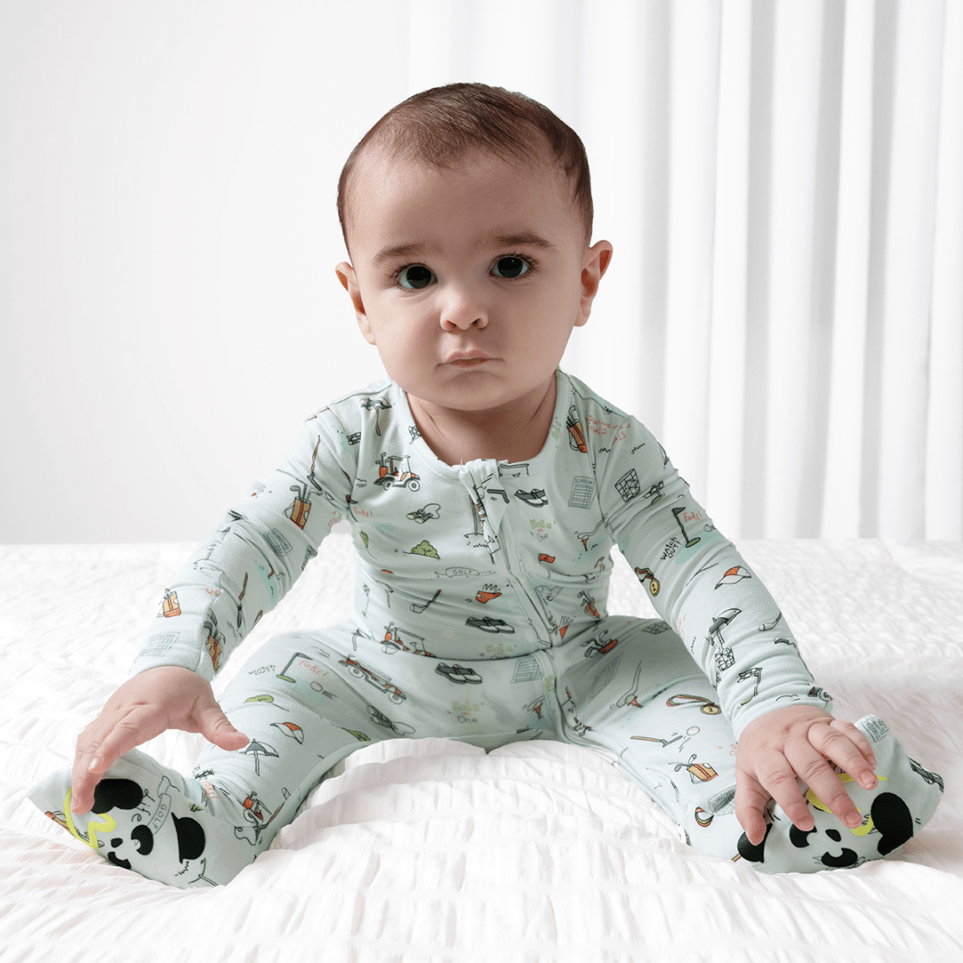 Baby wearing a golf patterned onesie sitting on a white surface with a white curtain in the background.