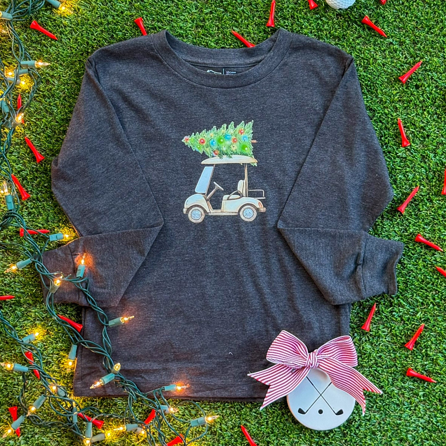 Charcoal long-sleeve kids’ shirt with a holiday golf cart graphic carrying a decorated Christmas tree, styled on turf with string lights, red golf tees, and a ceramic golf ornament.