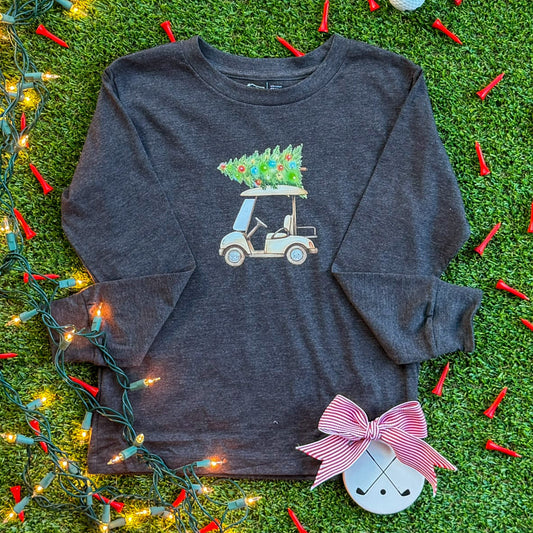 Charcoal long-sleeve kids’ shirt with a holiday golf cart graphic carrying a decorated Christmas tree, styled on turf with string lights, red golf tees, and a ceramic golf ornament.