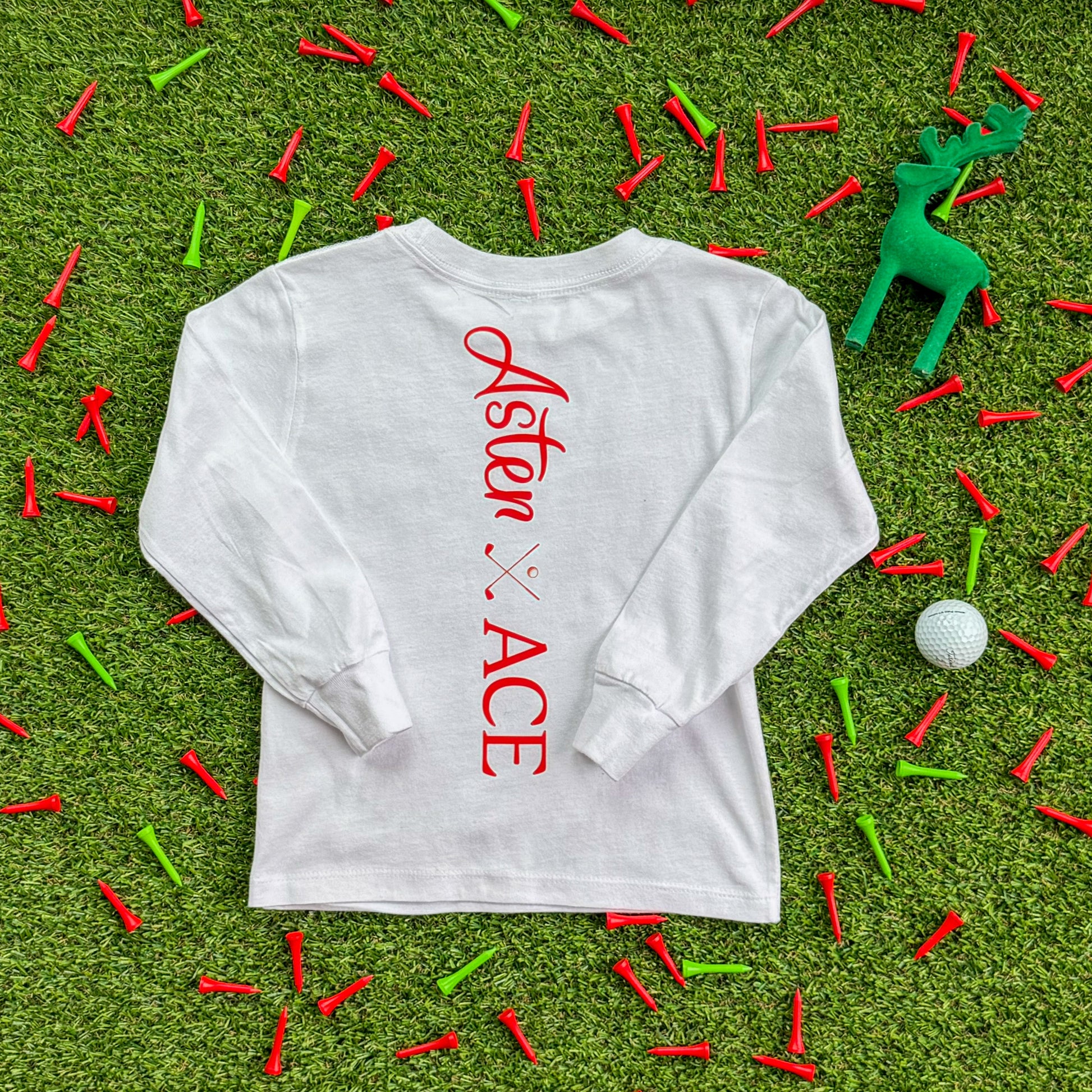Back view of a white long-sleeve kids’ shirt with a vertical red Aster and Ace logo printed down the spine, styled on turf with red and green golf tees and a green decorative reindeer.