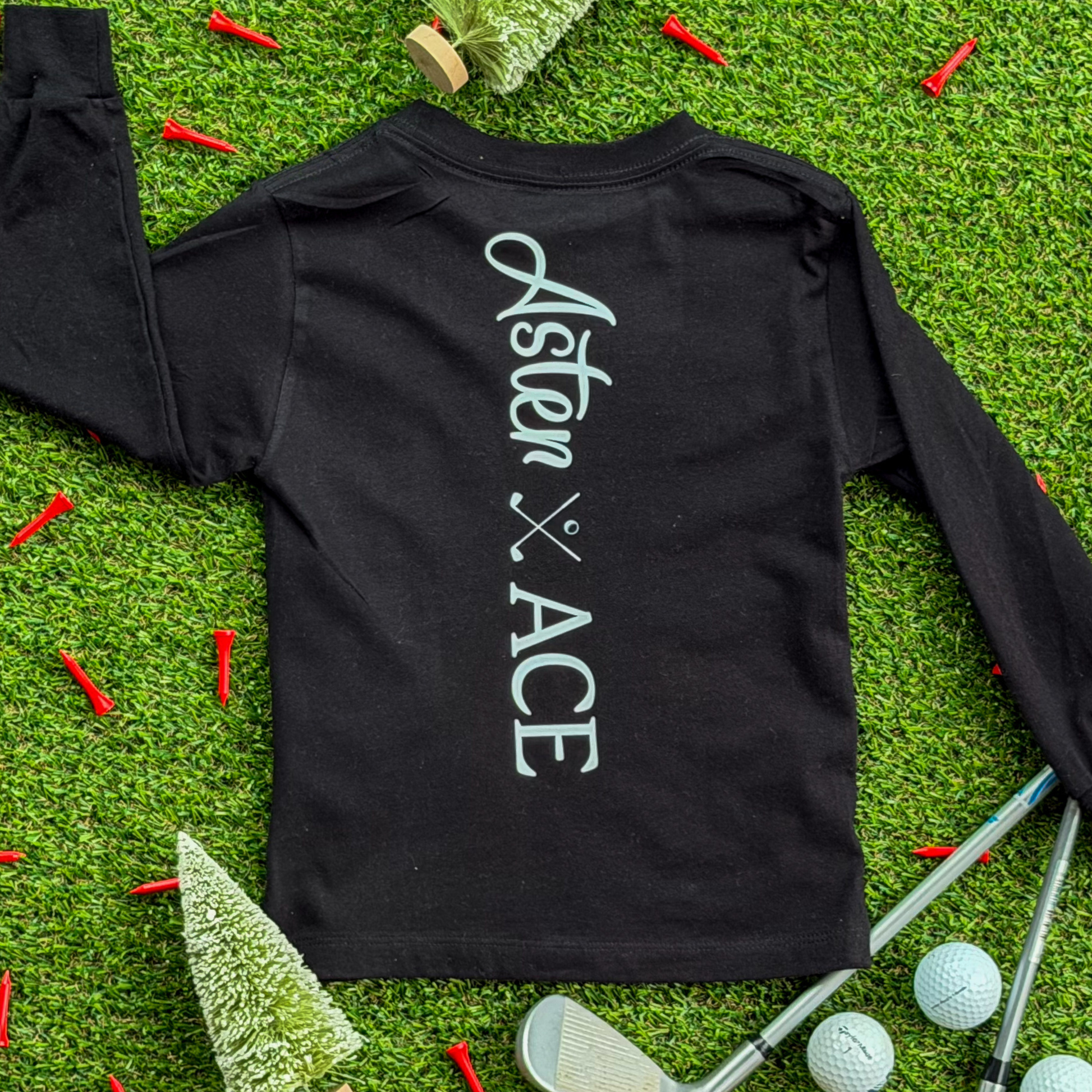 Back view of a black long-sleeve kids’ golf shirt featuring the Aster and Ace logo printed vertically in white, surrounded by golf balls, tees, and Christmas trees on green turf.