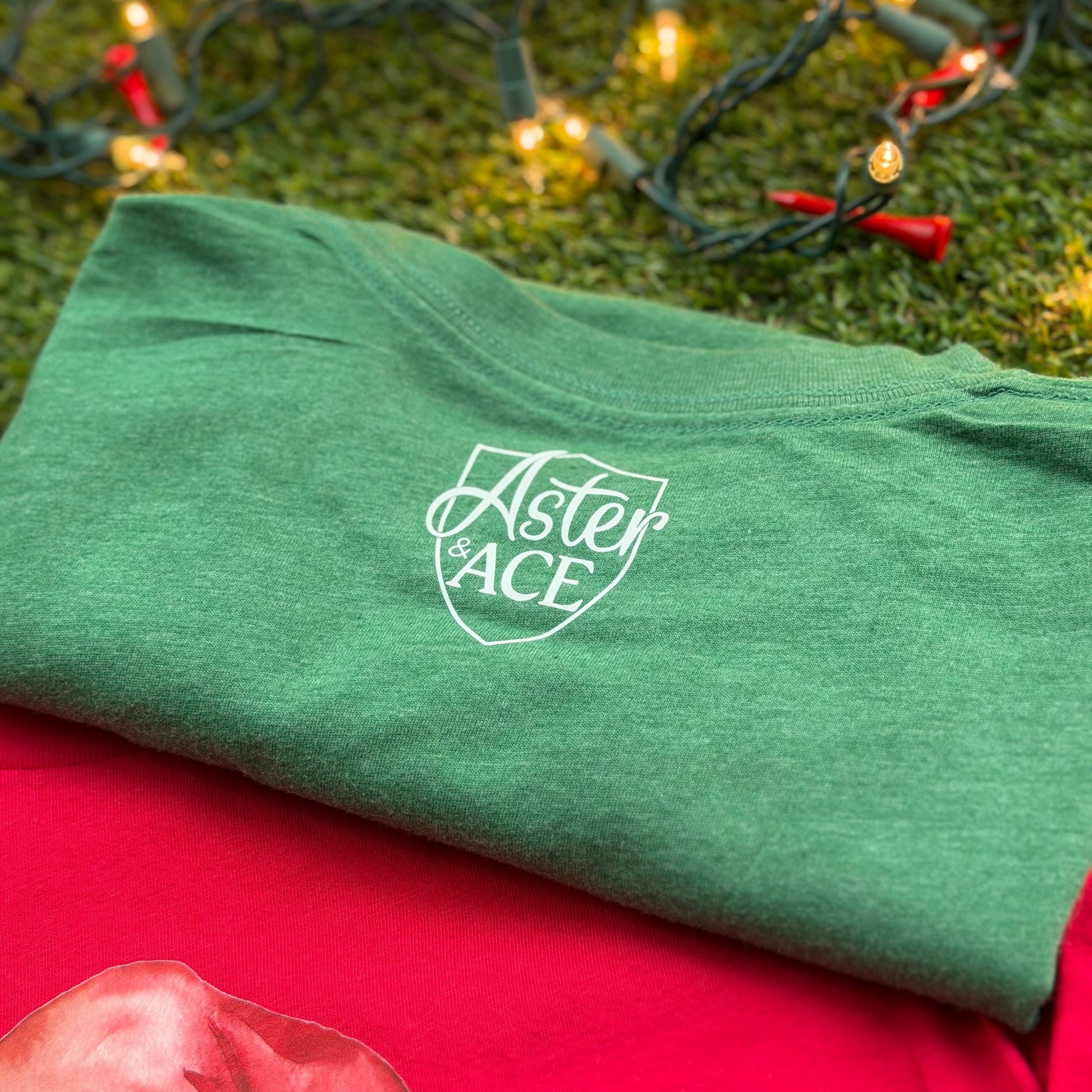 Folded heather green Aster and Ace t-shirt showing the back with white chest logo, displayed on turf with Christmas lights and red golf tees.