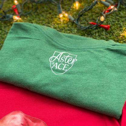 Folded heather green Aster and Ace t-shirt showing the back with white chest logo, displayed on turf with Christmas lights and red golf tees.