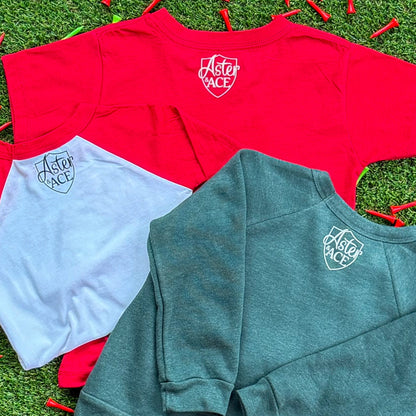 Back view of three Aster and Ace holiday tops—red short sleeve, red-and-white baseball tee, and heather green sweatshirt—showing the Aster and Ace logo printed on each, displayed on turf with scattered red golf tees.