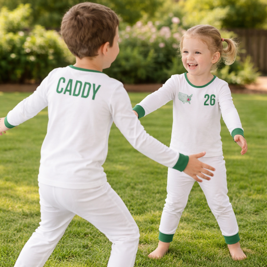 Two young children wearing matching white pajamas with green trim, one showing “CADDY” on the back and the other with a “26” graphic, playing together outdoors on grass