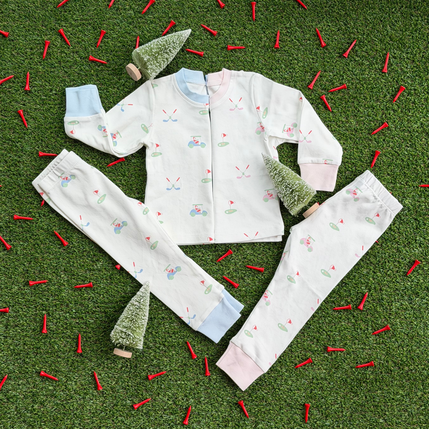 Two sets of kids’ Christmas golf pajamas laid flat on grass with mini holiday trees and red golf tees, showing matching tops and pants in pink and blue cuff styles with Santa golf print.