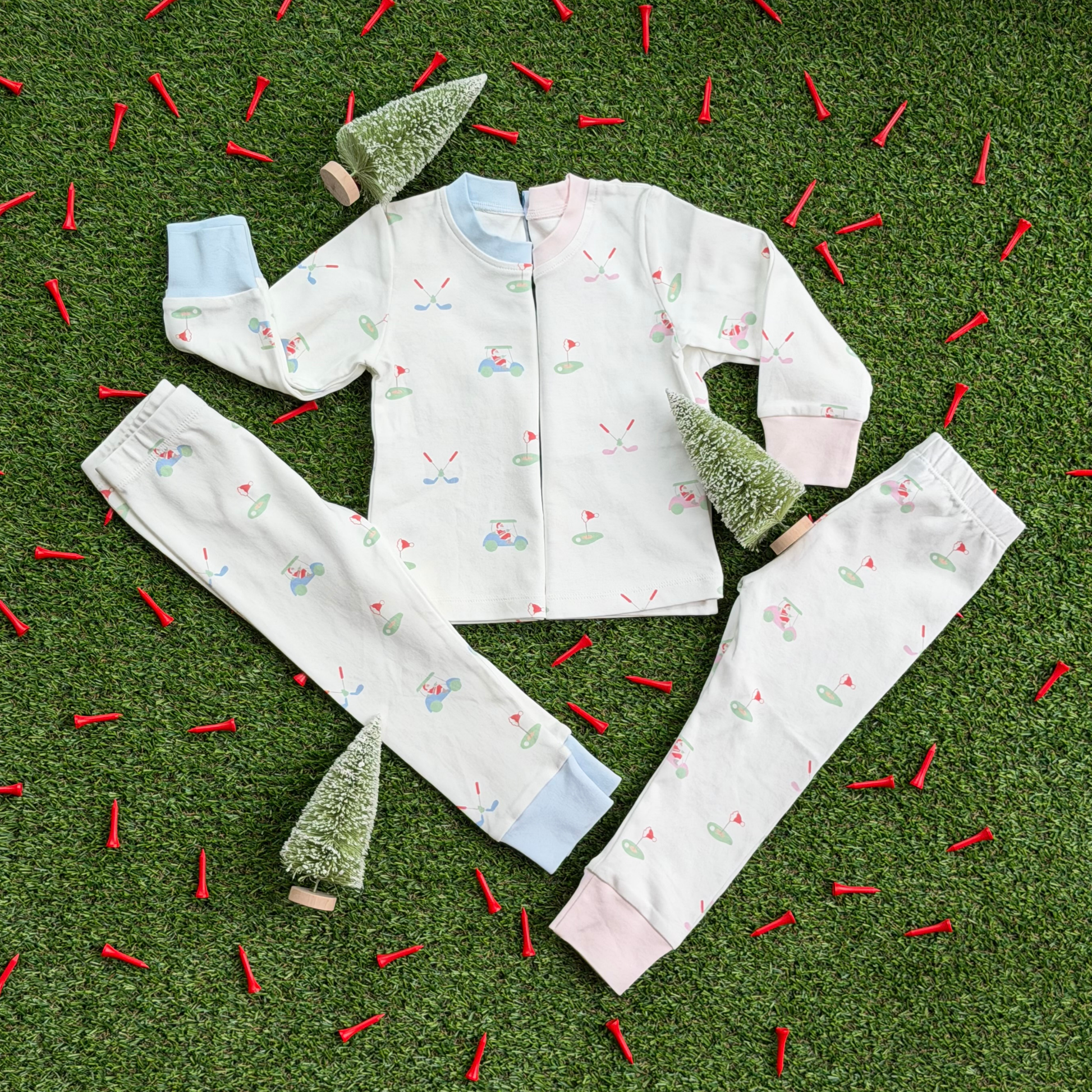 Two sets of kids’ Christmas golf pajamas laid flat on grass with mini holiday trees and red golf tees, showing matching tops and pants in pink and blue cuff styles with Santa golf print.