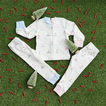 Two sets of kids’ Christmas golf pajamas laid flat on grass with mini holiday trees and red golf tees, showing matching tops and pants in pink and blue cuff styles with Santa golf print.
