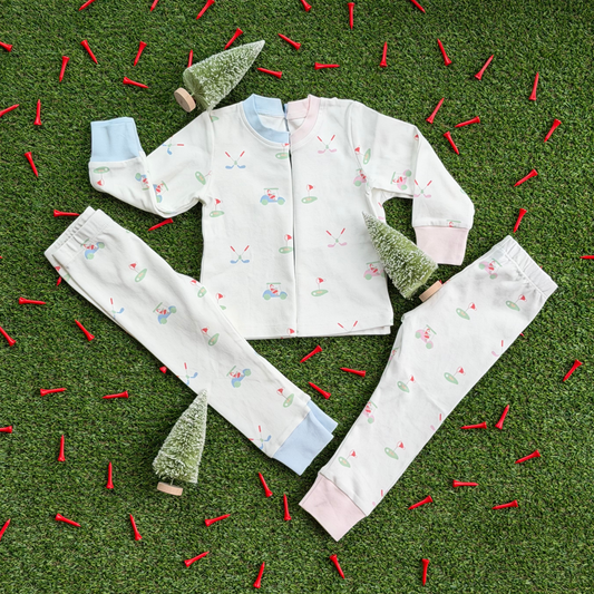 Two sets of kids’ Christmas golf pajamas laid flat on grass with mini holiday trees and red golf tees, showing matching tops and pants in pink and blue cuff styles with Santa golf print.