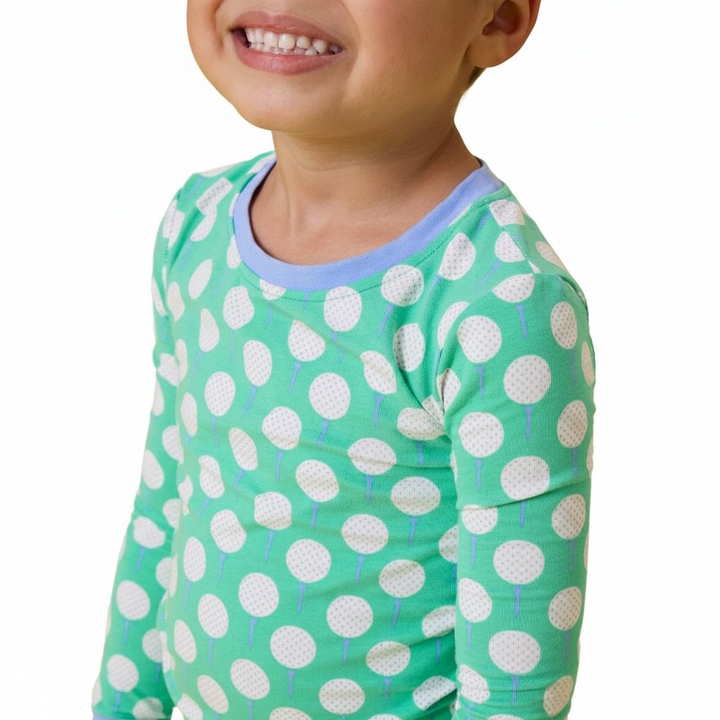 A close-up of a smiling child wearing a mint green long-sleeve top with white golf ball on tee pattern and light blue crew neck collar, against a plain white background.