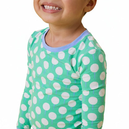 A close-up of a smiling child wearing a mint green long-sleeve top with white golf ball on tee pattern and light blue crew neck collar, against a plain white background.