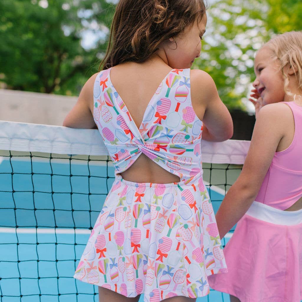 Young girl on a pickleball court wearing an activewear dress with a colorful pickleball paddle and bow pattern. Pictured from the back. Open back with a twist in the fabric.  