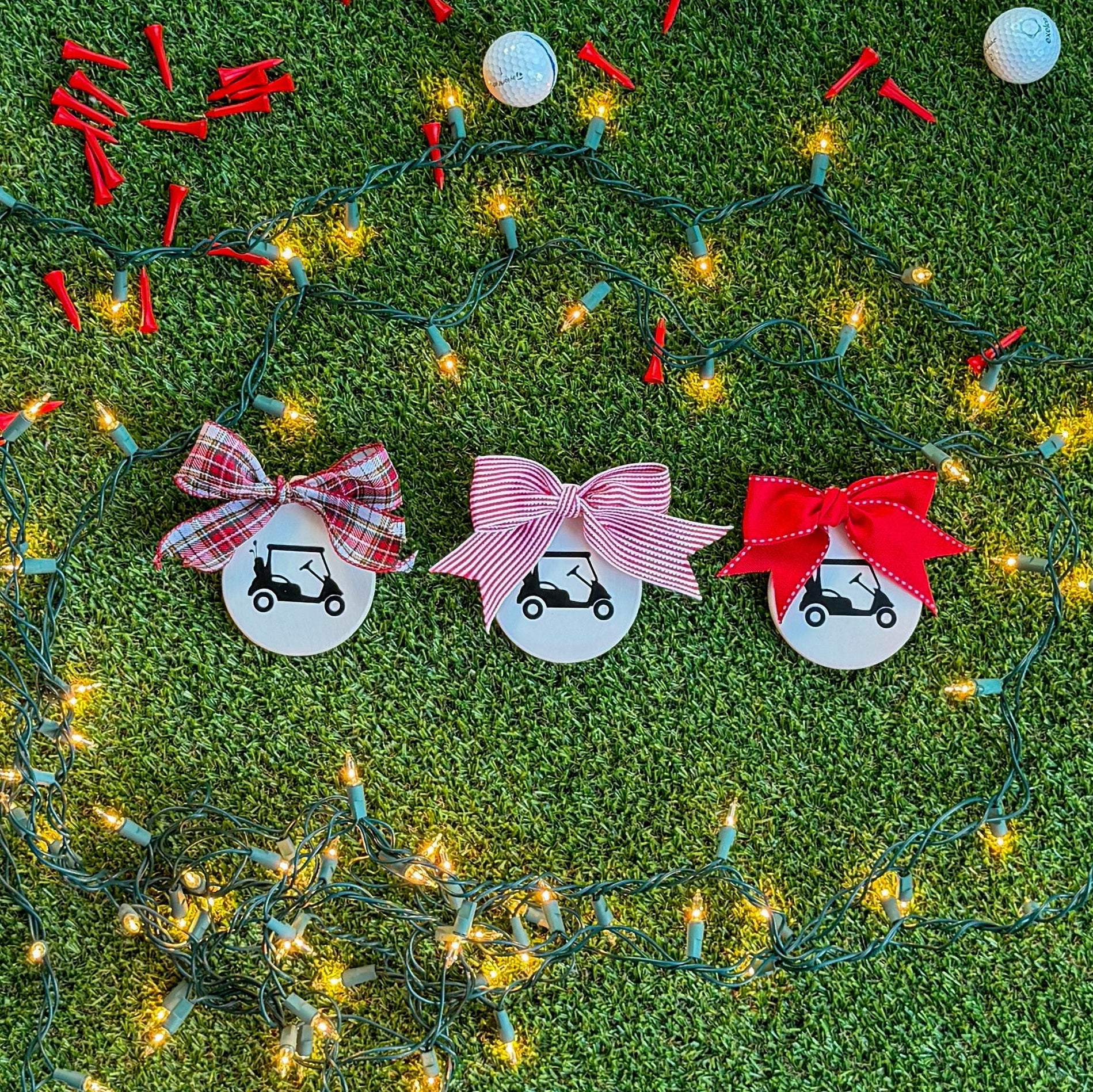 Three white ceramic ornaments featuring black golf cart silhouettes, each topped with a different ribbon — tartan, red-and-white stripe, and solid red — displayed on green turf with holiday lights and golf tees.