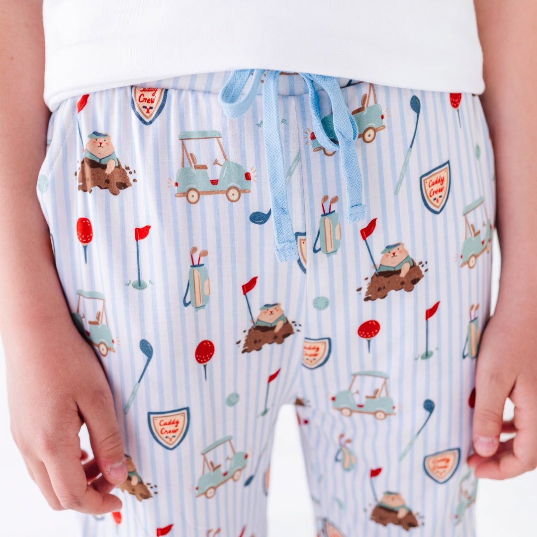 A close-up of a person wearing light blue striped pajama pants with a golf-themed print featuring golf carts, clubs, flags, and golf balls. The pants have a light blue drawstring waistband.