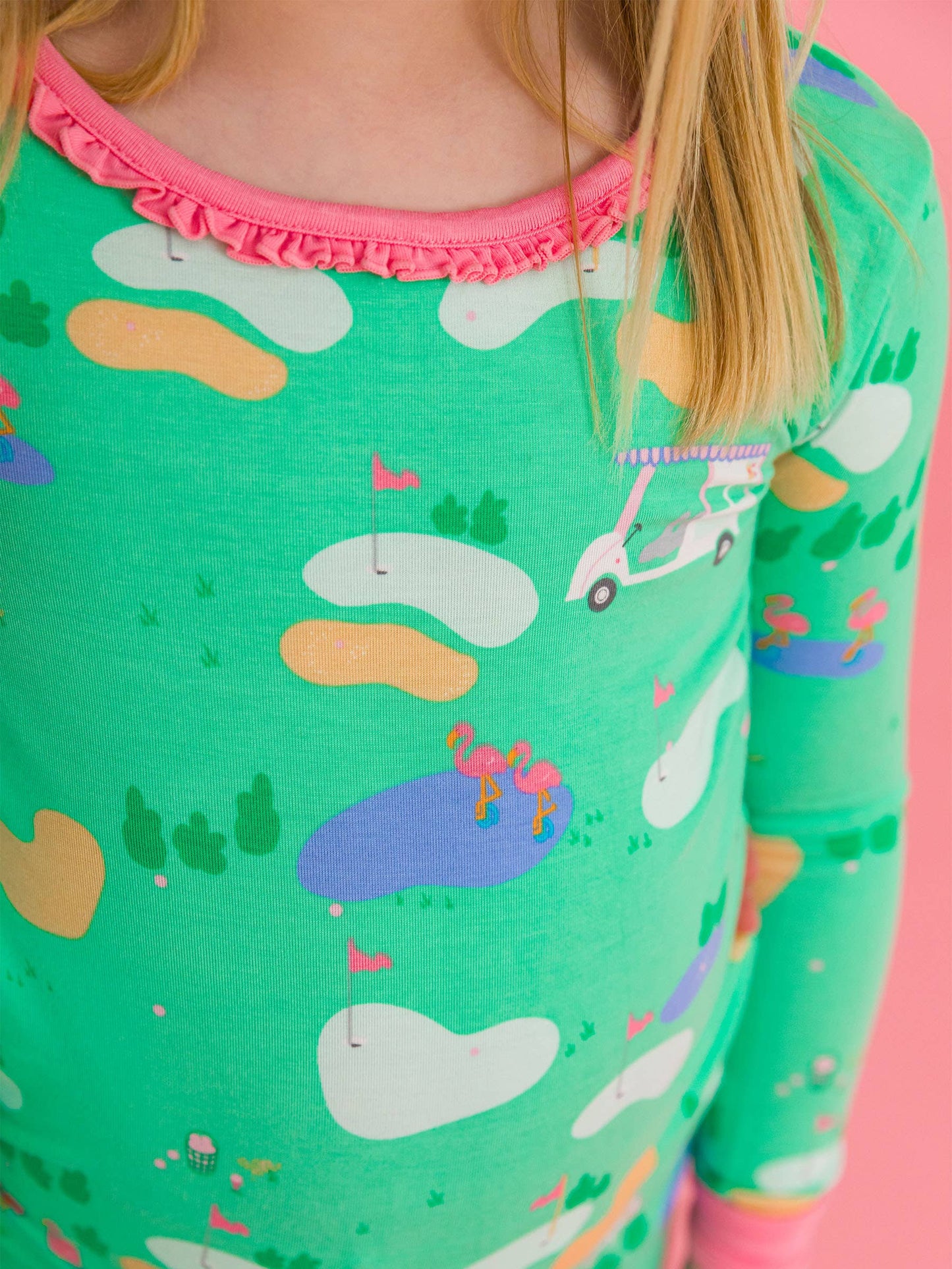 Close up shot of top of golf course pattern sleepwear. Green with blue water with flamingos, golf carts, bunkers, and greens with pink pin flags. Pink ruffle trim around the neck. 