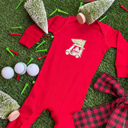 Red baby Christmas golf cart coverall with Santa driving and a decorated tree on top, displayed on turf with holiday décor, golf balls, tees, and small bottlebrush trees.