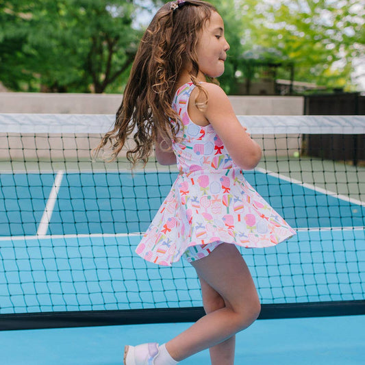 Young girl on a pickleball court wearing an activewear dress with a colorful pickleball paddle and bow pattern.
