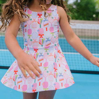 Young girl on a pickleball court wearing an activewear dress with a colorful pickleball paddle and bow pattern.