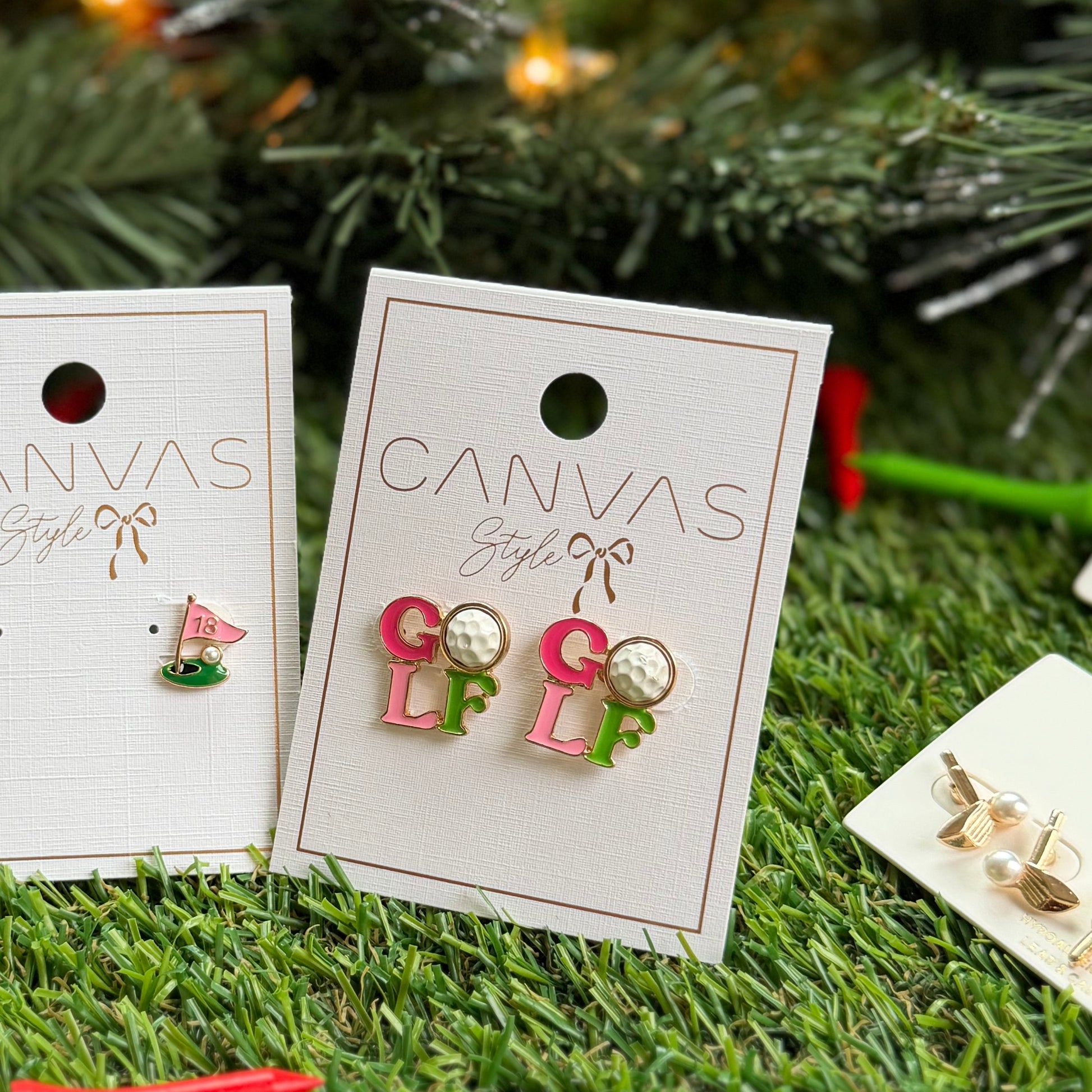 Golf-themed stud earrings from Canvas Style displayed on product cards, including colorful “GOLF” letter earrings with golf ball details and a pink 18th hole flag earring, photographed on artificial grass with a Christmas tree background for a festive gift look.