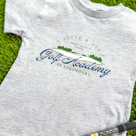 Aster and Ace Golf Academy kids graphic tee in heather grey styled on green turf with golf club detail.