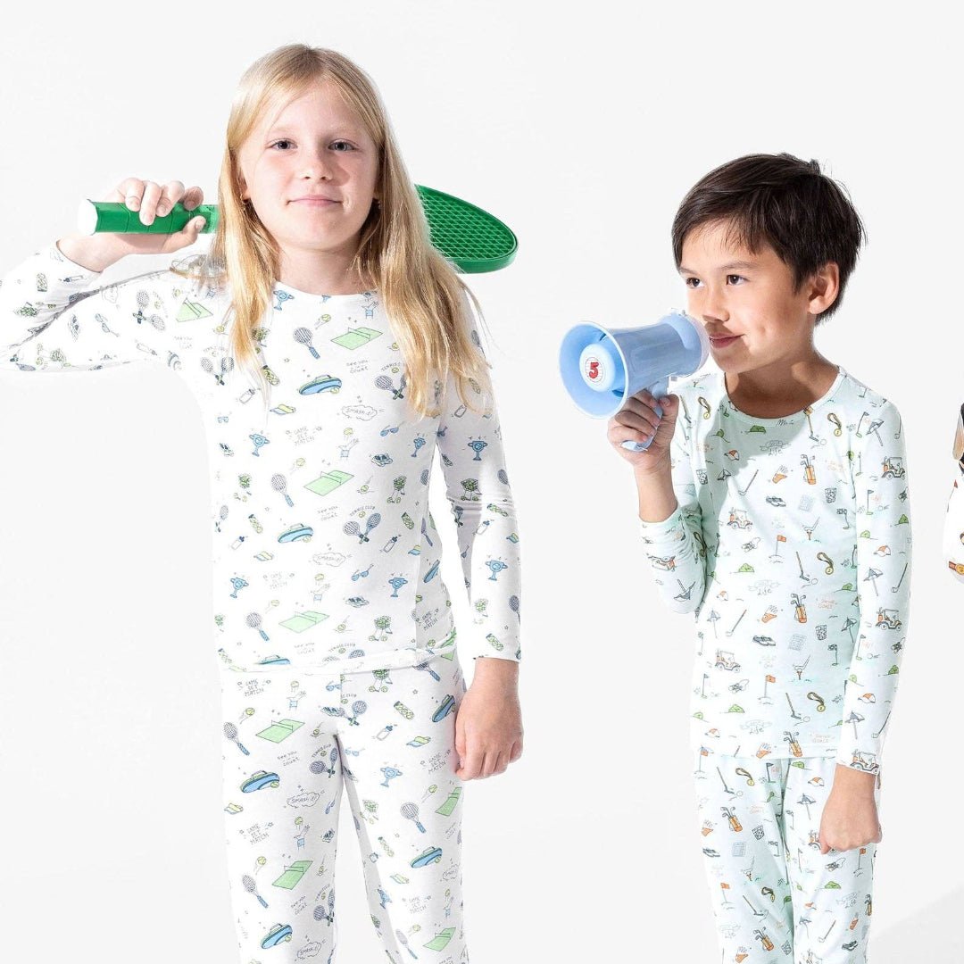 2 kids wearing pajamas. One has white pajamas with tennis icon doodles and the other has light blue pajamas with golf icon doodles.