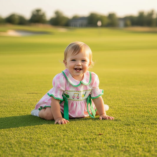 Baby girl sitting on a golf course wearing a pink smocked golf-themed bloomer set with green bow details