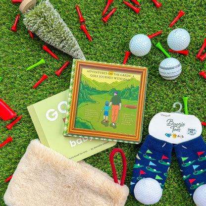 Kids’ golf-themed stocking stuffer gifts displayed on turf, featuring “Adventures on the Green: A Golf Journey With Dad” children’s book, “Golf Baby” board book, fuzzy golf ball zip pouch, and Boogie Toes rattle socks with golf flag designs surrounded by red and green golf tees and golf balls.
