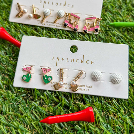 Golf-themed stud earring sets from Influence New York displayed on grass turf, featuring golf cart earrings, golf ball studs, pearl-accented clubs, and pink and green flag earrings, styled with red and green golf tees for a holiday gift look.