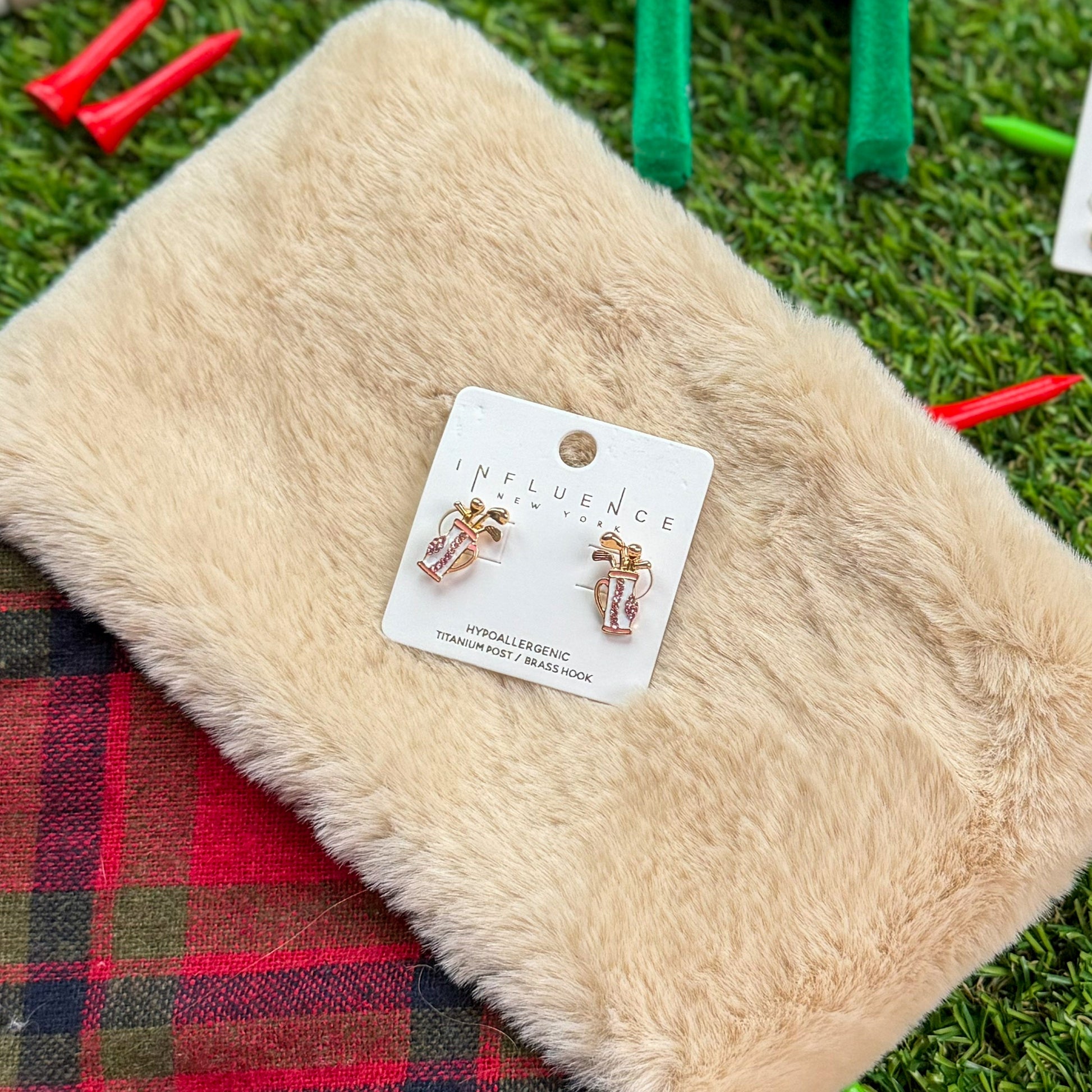 Golf-themed stocking stuffer flat lay featuring Influence New York pink golf bag stud earrings displayed on a fuzzy cream pouch with red plaid trim, surrounded by green turf and red golf tees.