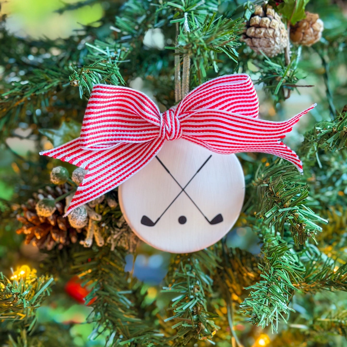 White ceramic ornament with a black crossed golf clubs silhouette, topped with a red-and-white striped ribbon, hanging on a Christmas tree with lights and pine cones.