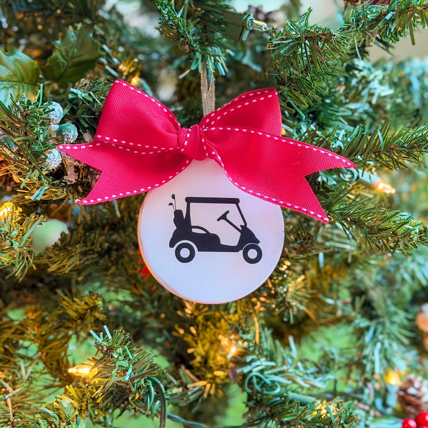 White ceramic ornament featuring a black golf cart silhouette, tied with a large red stitched ribbon and hanging on a decorated Christmas tree.