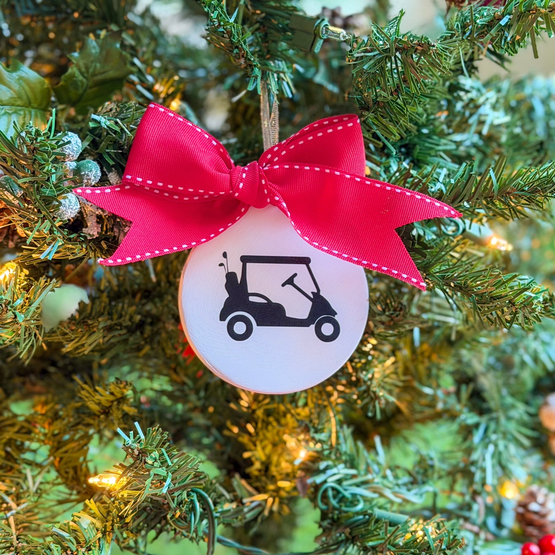 White ceramic ornament featuring a black golf cart silhouette, tied with a large red stitched ribbon and hanging on a decorated Christmas tree.