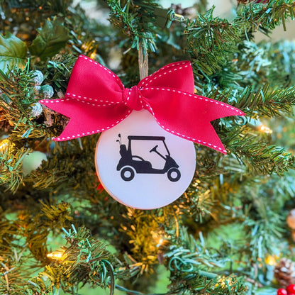 White ceramic ornament featuring a black golf cart silhouette, tied with a large red stitched ribbon and hanging on a decorated Christmas tree.