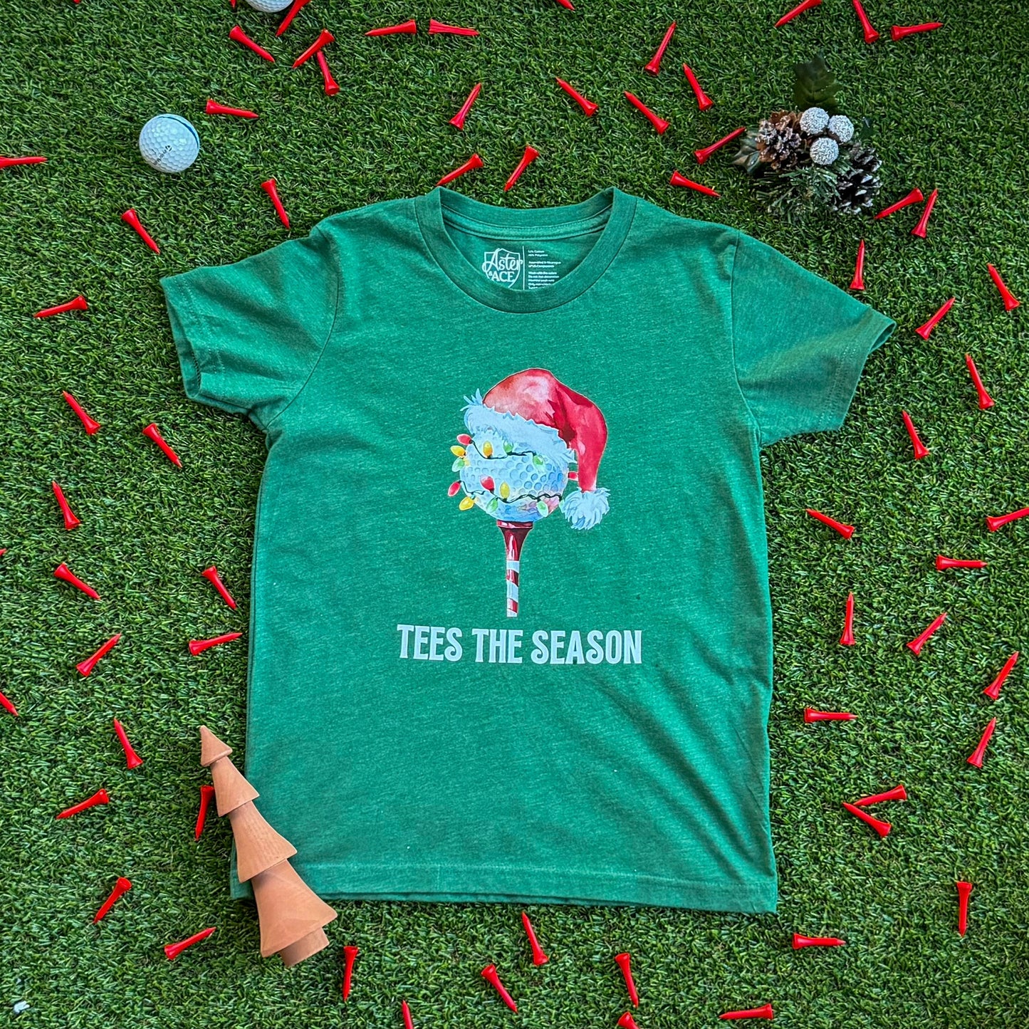 Heather green kids’ Christmas golf shirt featuring a Santa hat golf ball on a tee and “Tees the Season” text, laid flat on turf with scattered red golf tees and holiday decorations.