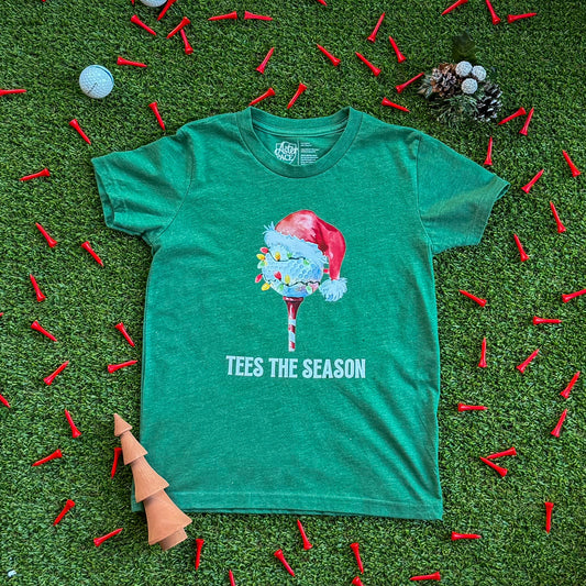Heather green kids’ Christmas golf shirt featuring a Santa hat golf ball on a tee and “Tees the Season” text, laid flat on turf with scattered red golf tees and holiday decorations.