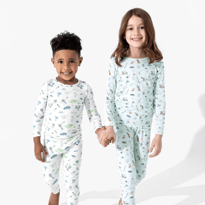 Two children modeling matching long-sleeve pajama sets against a clean white studio background. The boy wears a white pajama set with a small multicolor tennis print, and the girl wears a light blue pajama set with a small multicolor golf print.