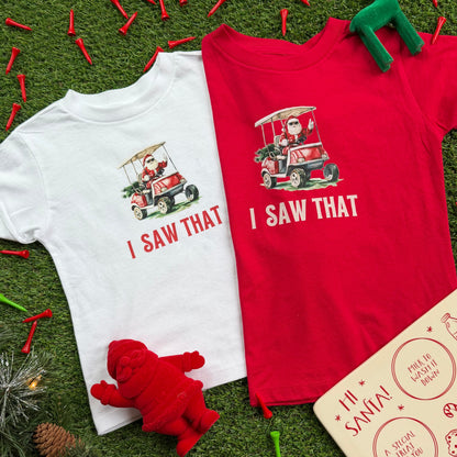 White and red kids’ t-shirts featuring a Santa golf cart graphic and the text “I Saw That,” displayed on turf with holiday decor, golf tees, and a plush red Santa figurine.