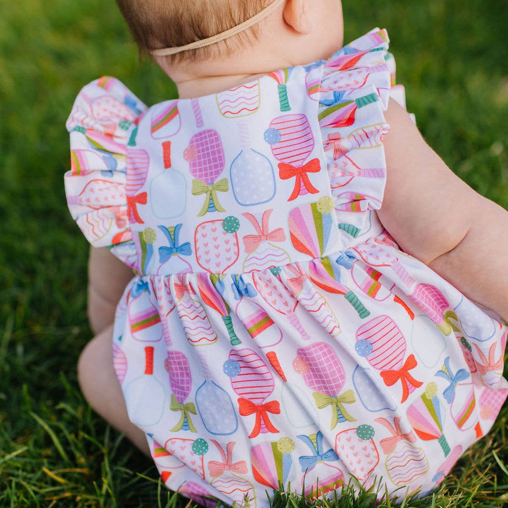 Baby sitting in grass wearing ruffle-sleeved romper with a colorful bow and pickleball paddle pattern. 