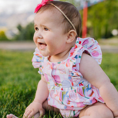 Close up shot of baby sitting in the grass with ruffle-sleeved romper that has a colorful pickleball paddle and bow pattern. The top of the romper also has two buttons.