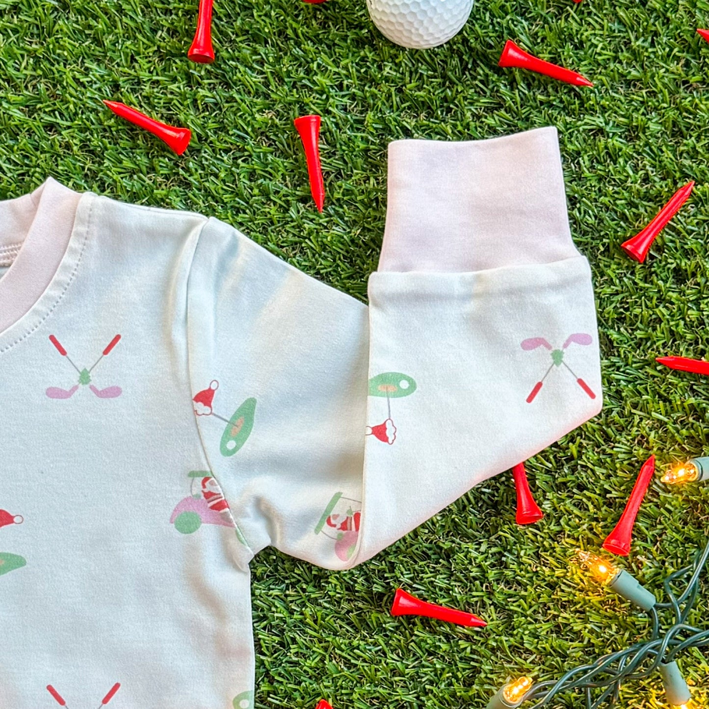Close-up of a white children’s pajama sleeve featuring pastel holiday golf-themed illustrations, including golf clubs, Santa hats, and greens, displayed on turf with red golf tees and Christmas lights.
