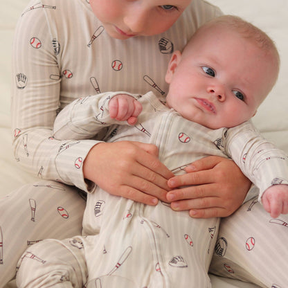 Baby and big kid wearing matching pajamas with neutral stripes, baseball bats, baseballs, and gloves on them.